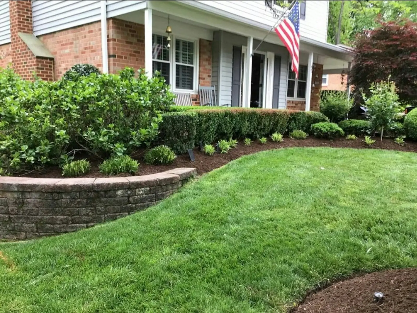 Brick house front yard with mulched shrub beds, a stone retaining wall and a green lawn